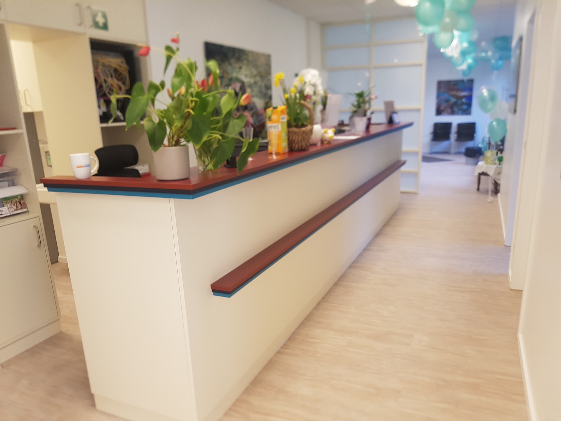 Bild einer Praxis mit Holzm&ouml;beln und Holzboden. Zu sehen ist ein langer Tisch, auf dem viele Blumen stehen. Im Hintergrund sind blaue Ballons an der Decke zu sehen. 
