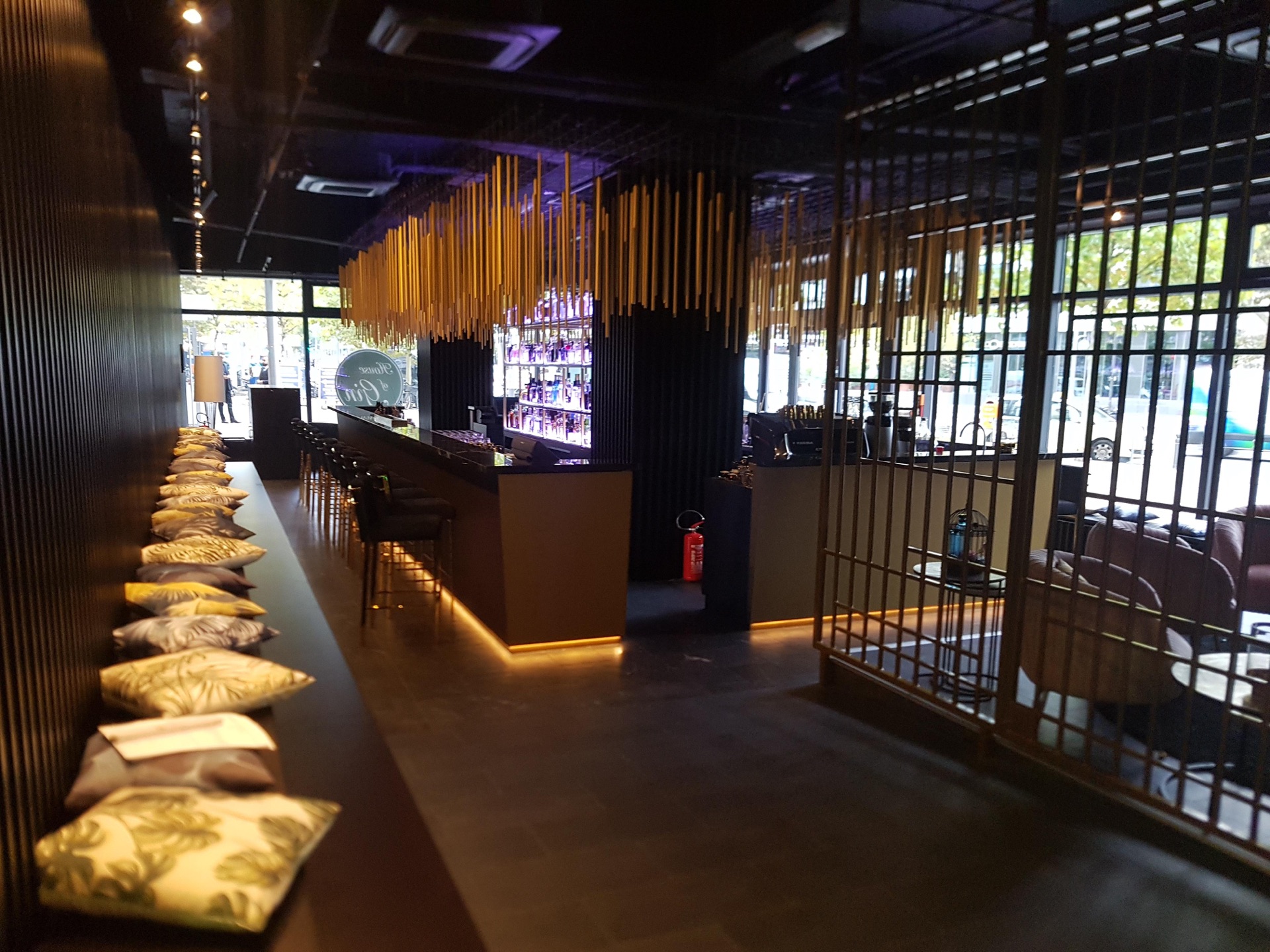 Bild einer Bar mit gem&uuml;tlicher Einrichtung. Braunes Holz und gelbes Licht unten an den Tischen stechen hervor. Im Zentrum sind regale mit Gl&auml;sern und drum herum Barhocker am eckigen Tisch. 