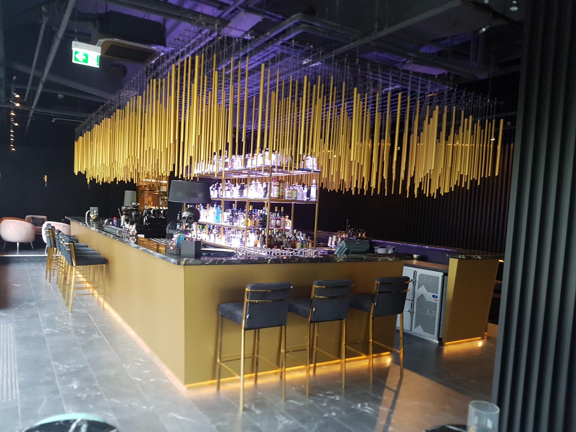 Bild einer Bar, die von einem eckigen, gelben Tisch umgeben ist. In der Mitte ist ein Regal mit zahlreichen Flaschen.