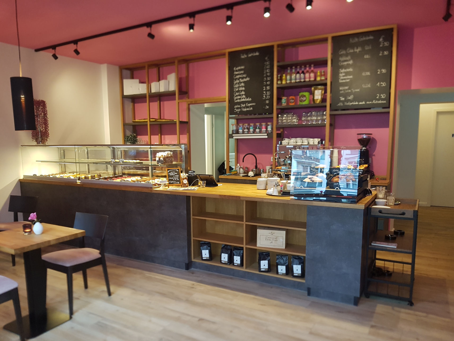Bild eines Café's. H&ouml;lzerner Tresen, pinke Wand mit Regalen voller Flaschen und zwei Angenotstafeln. Auf dem Tresen sind Vitrinen mit Backwaren.